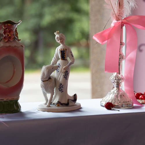 Kompozycja złożona z porcelanowej figurki kobiety z psem, ozdobnego wazonu, świecznika z różową wstążką i dekoracyjnych wiśni na białym obrusie.
