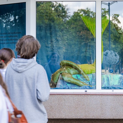 Publiczność ogląda przez szybę instalację artystyczną z zielonym ogonem syreny oraz nadrukowanym tekstem.