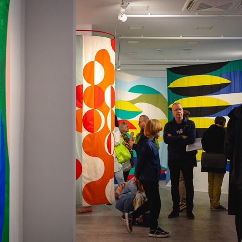 Odwiedzający spacerują po galerii pełnej barwnych tkanin w abstrakcyjne, geometryczne wzory.