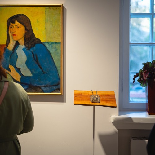 Zwiedzający oglądają obraz kobiety w niebieskim stroju oraz ozdobną biżuterię wystawioną obok okna galerii.