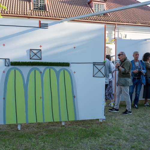 Grupa osób zwiedza wystawę plenerową, oglądając kolorowe, geometryczne obrazy.