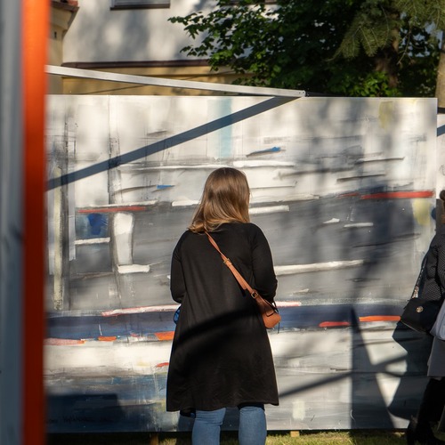 Kobieta w czarnej bluzce ogląda abstrakcyjny obraz podczas wystawy plenerowej w słoneczny dzień.