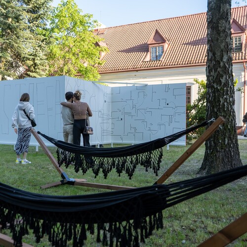 Ludzie oglądają minimalistyczne rysunki na białych tablicach podczas wernisażu, obok rozwieszonych hamaków na trawniku.