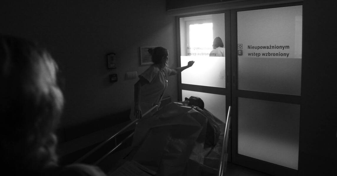 Czarno - biała fotografia reportażowa. Pielęgniarka w szpitalu wiezie pacjenta  na łóżku szpitalnym przez drzwi z napisem „Nieupoważnionym wstęp wzbroniony”.