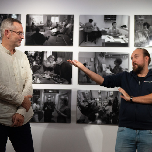 Autorzy wystawy rozmawiają na tle ściany z czarno-białymi fotografiami dokumentującymi codzienne życie w szpitalu.