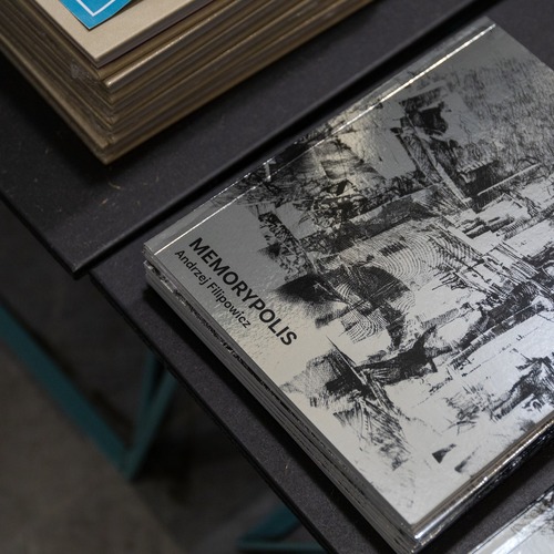 Stos katalogów wystawy „MEMORYPOLIS” autorstwa Andrzeja Filipowicza z okładką przedstawiającą czarno-biały rysunek architektury.