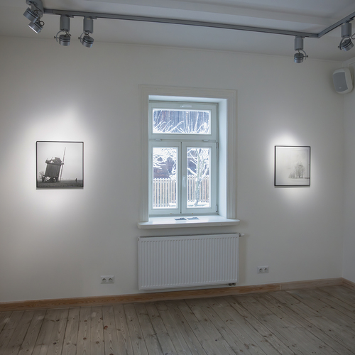 Wnętrze galerii z czarno-białymi fotografiami zimowego krajobrazu zawieszonymi na białych ścianach wokół okna z widokiem na zaśnieżone podwórze.