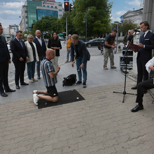Mężczyzna klęczy na chodniku w centrum miasta, otoczony przez grupę osób, podczas gdy obok gra akordeonista, a inny mężczyzna przemawia do mikrofonu.